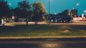 Ciemata autobusa pietura un satiksme vakarā, izgaismota ar laternām Latvijas pilsētas vidē.