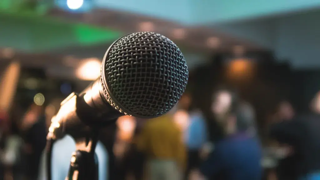 Tuvplānā redzams melns vokālais mikrofons uz statīva ar izplūdušu auditoriju fona.