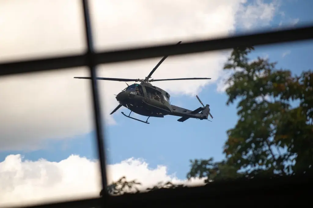 Militārais helikopters lido debesīs, skatoties caur loga rāmja stiklojumu ar redzamiem kokiem.