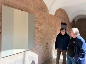 Divi vīrieši ar brillēm stāv mākslas galerijā pie liela, minimālistiska, gaišas krāsas gleznojuma.