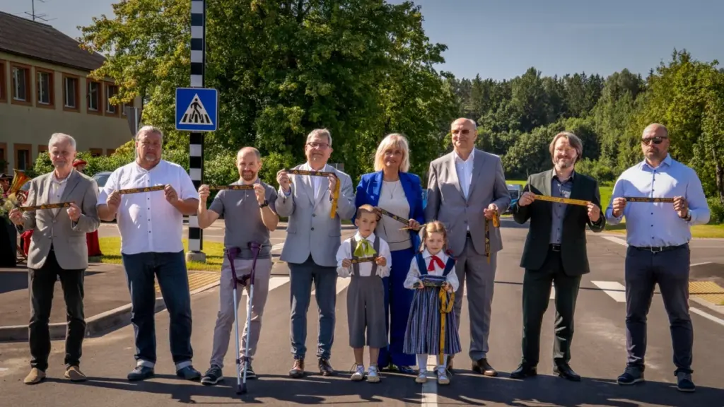 Grupa cilvēku un bērni uz jaunas asfalta ielas svinīgi pārgriež simbolisku dzeltenu lenti.