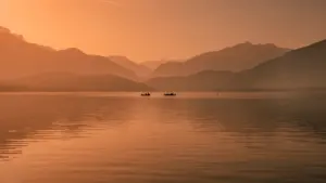 Mierīgs ezers rītausmas gaismā ar divām nelielām laivām tālplānā.