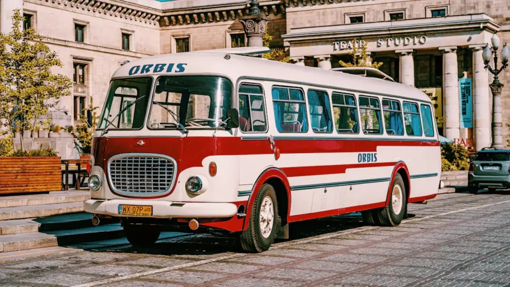 Sarkans un balts autobuss stāv pilsētas laukumā pie vēsturiskas ēkas.