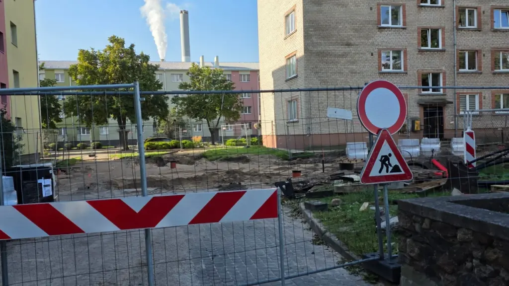 Norobežots būvlaukums pilsētvidē ar ceļa zīmi, kas brīdina par notiekošiem remontdarbiem pie daudzdzīvokļu mājām.