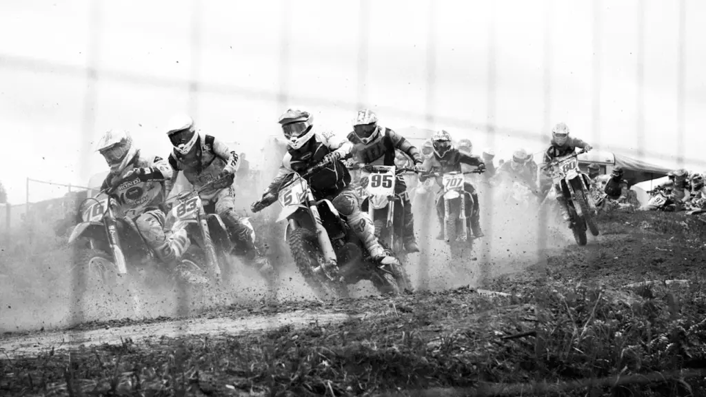 Motokrosa sacensību dalībnieki starta brīdī, braucot pa trasi melnbaltā fotogrāfijā.
